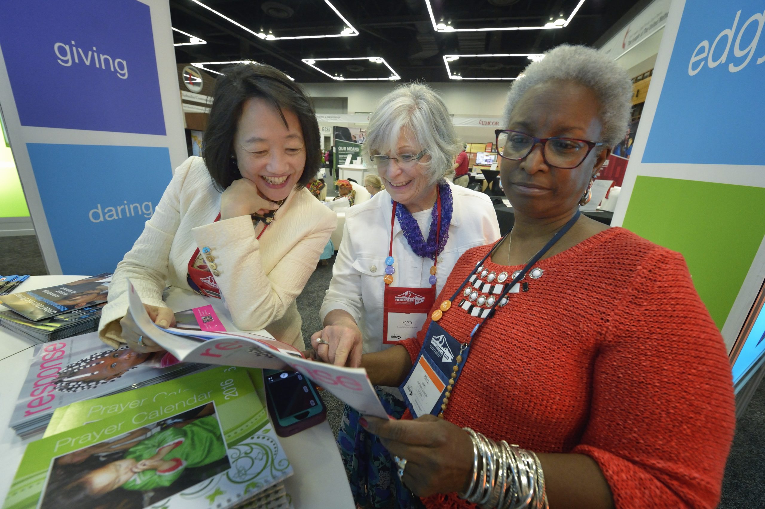 Three women read a magazine together standing at a colorful display in an exhibit hall.